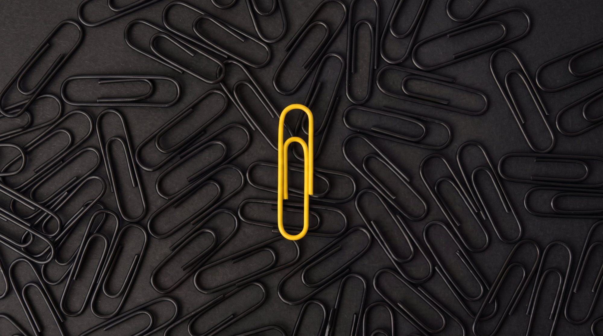 Um clipe amarelo em pé no centro de dezenas de clipes pretos espalhados sobre uma superfície escura